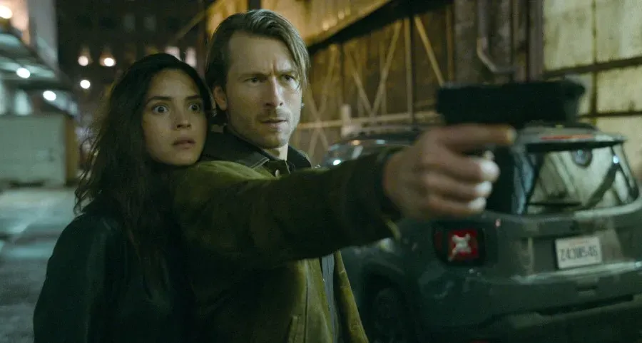 Adria Arjona standing behind Glen Powell, who is pointing a gun, in the film Hit Man
